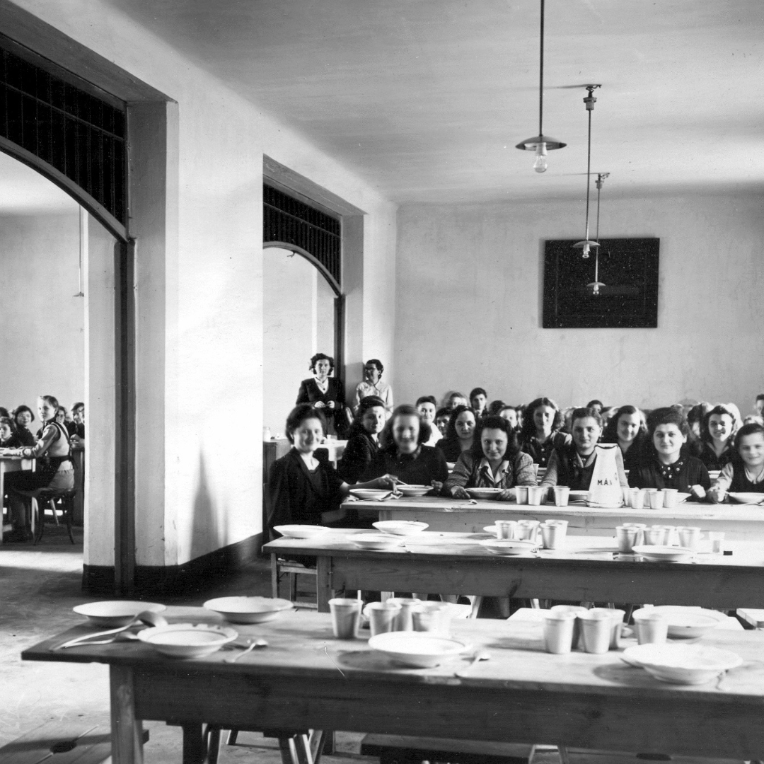 Children and a number of adults sitting in the canteen of an orphanage in Hungary in 1926. The first two rows of the tables are empty. The left side of the room is divided off by a wall with two large doors open.