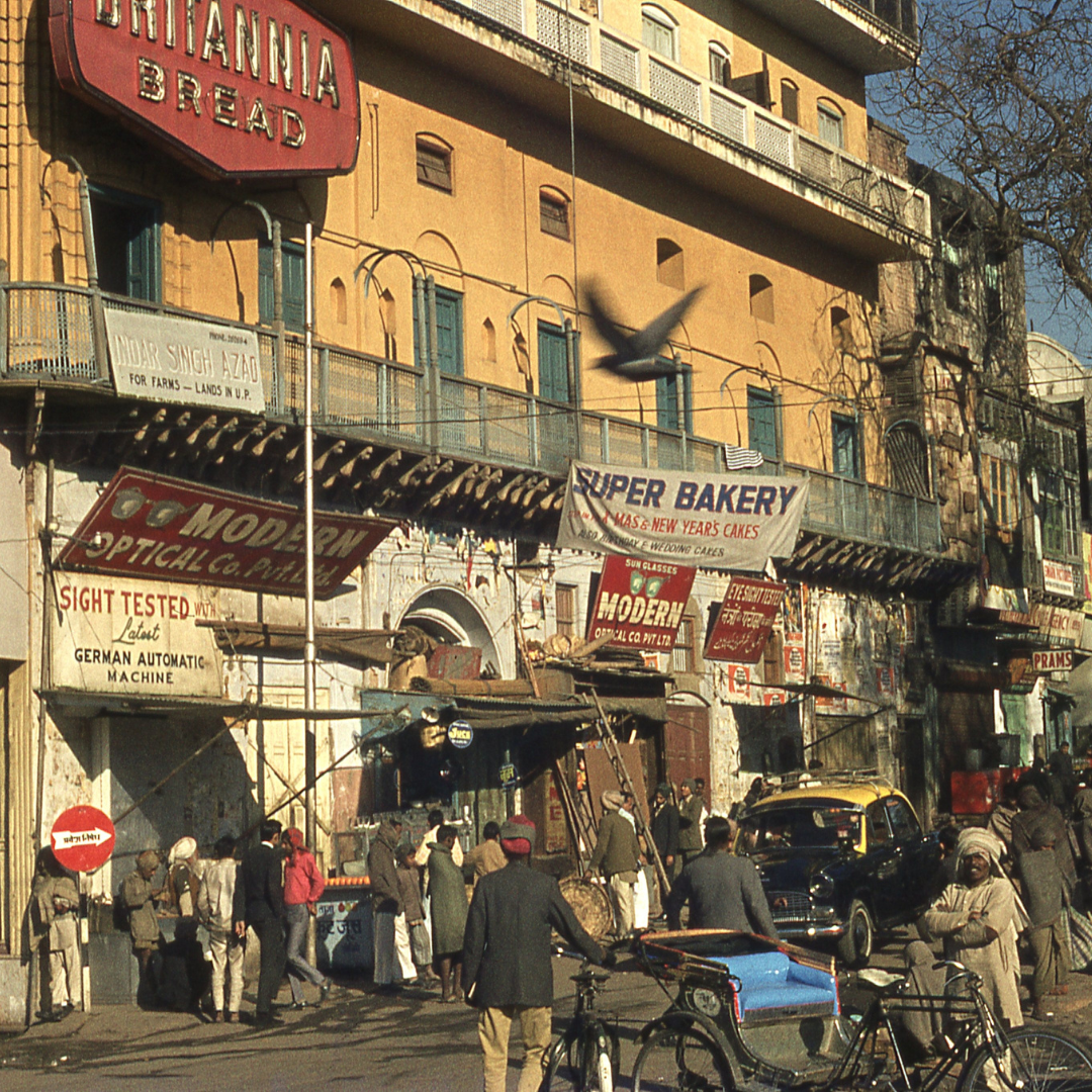 Busy street in 1960s India. Colour picture. People in front of a large yellow building with various shop signs and advertising signs.