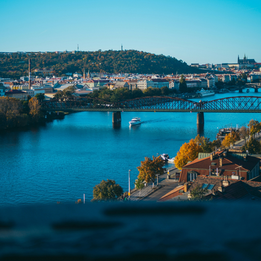 View of Prague from Vyšehrad. The Vltava river runs through the middle of the picture and it is crossed by a bridge. There is a small ship passing under the bridge. Prague Castle is visible in the distance on the right hand side. The left hand side is dominated by Petrin hill.
