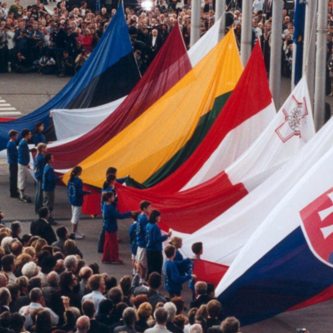 The flags of East European countries being hoisted during the celebrations of European Union enlargement in 2024. Children are holding up the flags. People are standing around.