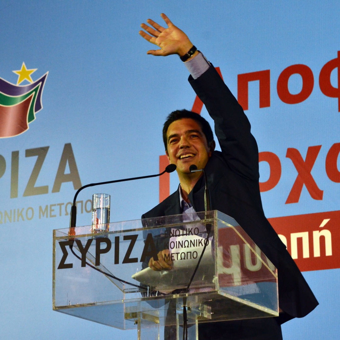 Alexis Tsipras, leader of the left-wing political party SYRIZA at a 2012 election rally. He is in the centre of the photo behind a see-through lectern that has the party's name on it. He is waving with his left arm and smiling. He is dressed in black suite. Behind him there is a light blue backing with partially visible Greek text. On the left-hand side the party's logo is visible on the wall.