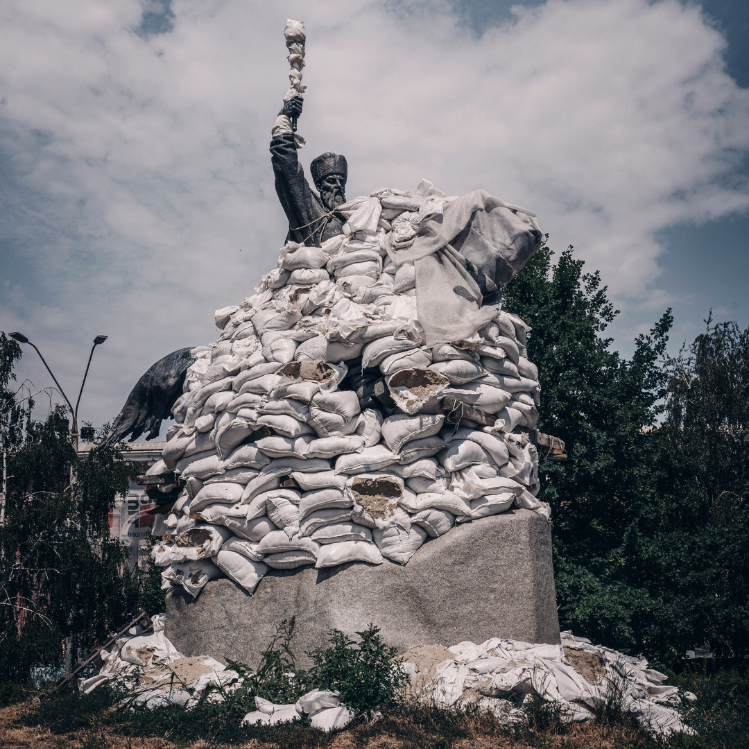 Monument of Petro Sahaidachnyi in Kyiv. The statue is covered with sandbags, only the statue's head and raised left arm are visible. There are trees and vegetation around the statue. The background has a muted blue sky with white clouds.