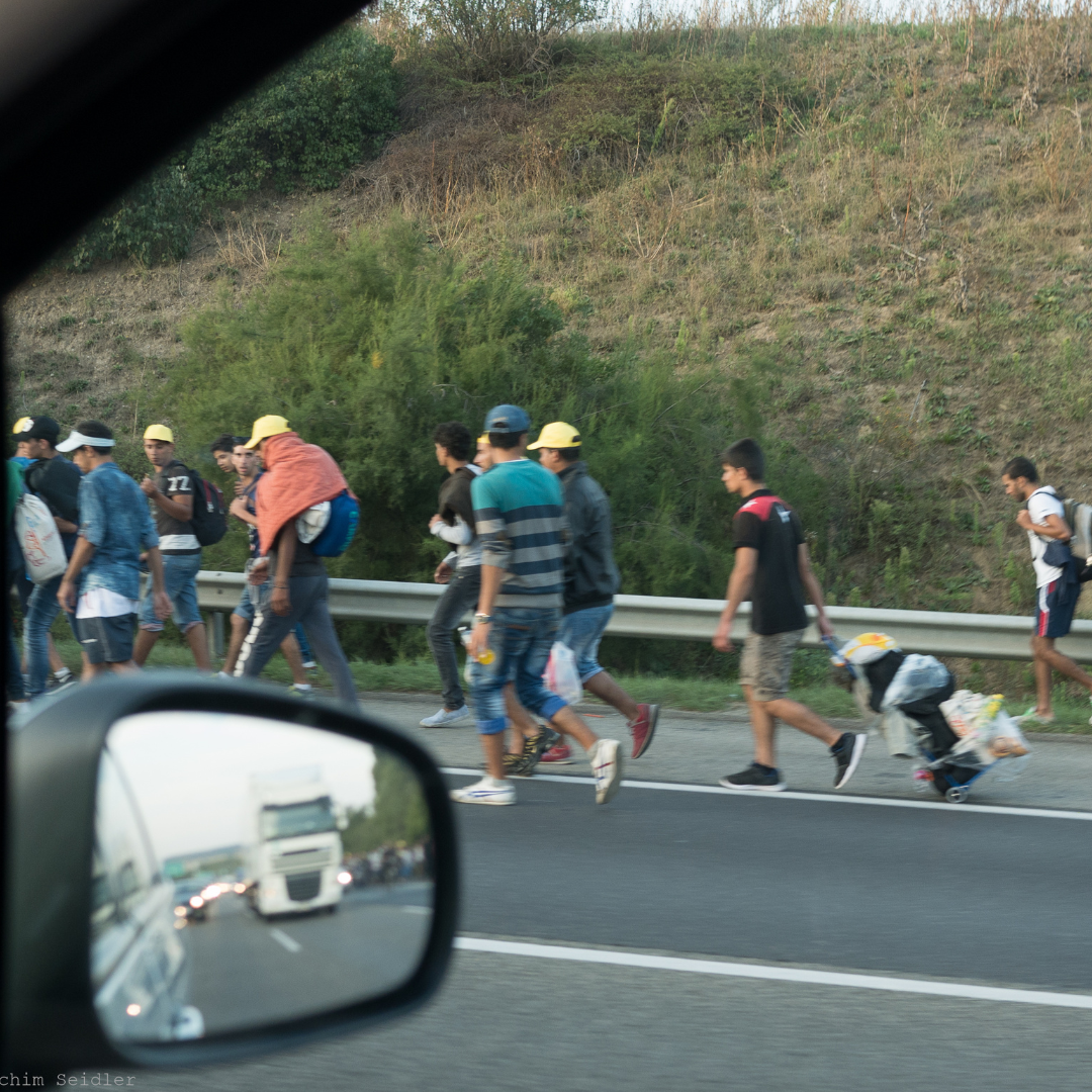 Refugees, mostly young men, some with suitcases walking on the shoulder of a motorway in Hungary. The picture was taken from inside a car. The mirror of the car is shown in the bottom left corner. In the mirror a white truck is reflected.