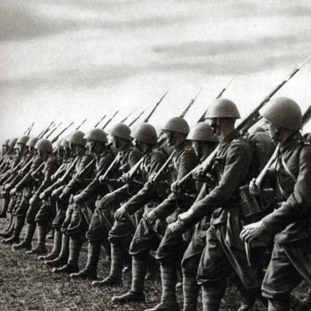 Black and white photo of soldiers marching in line, with riffles on their shoulders. They are wearing combat gear hats and are looking to their right, so their faces are not visible on the photo. They seem to be marching on a field, on the left had corner some grass is visible. The sky seems overcast, with clouds.