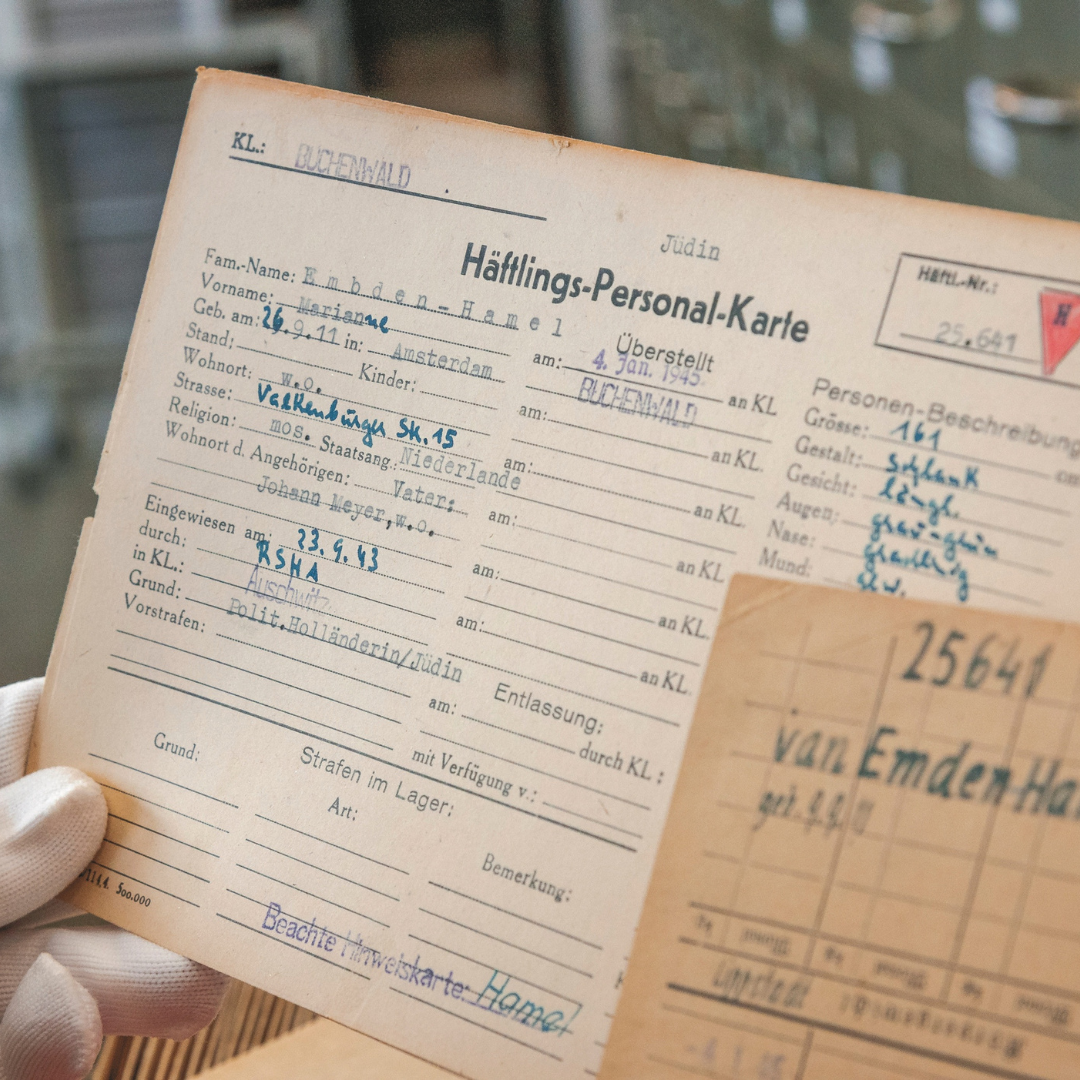 A gloved hand holding a card with German writing on it. The card is an inmate information card from Buchenwald. There is another, smaller card in the right hand corner of the photo with numbers listed on it. The larger card looks light brown with time, the smaller card is a darker shade of brown.