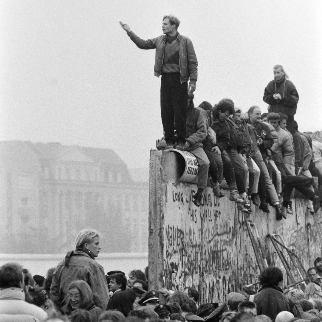 Black and white photo of the fall of the Berlin Wall. A man standing on top of the destroyed Berlin Wall, seemingly pointing into the distance. There is a crowd sitting behind him on the wall and there is another crown standing around the wall.