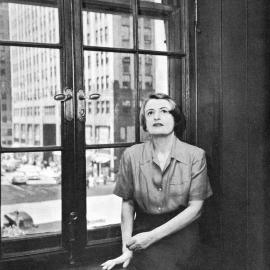 Ayn Rand sitting on a window sill with a New York daily life in the background, including a building with windows and a taxi. She is wearing a short-sleeved blouse and what appears to be a black skirt.