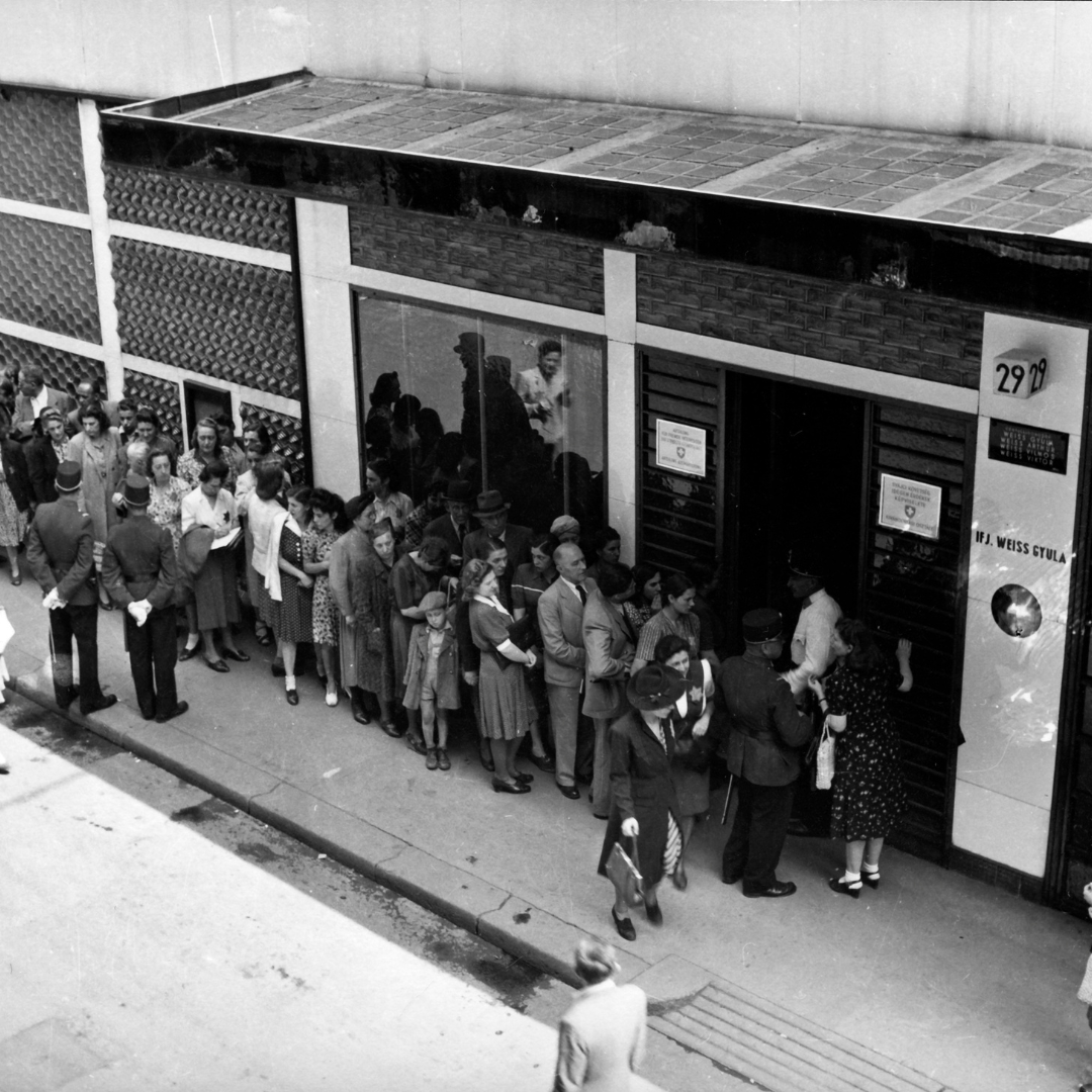 People queuing in front of the Swiss Embassy’s Emigration Department for Representing Foreign Interest in Budapest in 1944. The picture is black and white. The entrance of the building has a glass roof and from what is visible from the front also has large windows. The rest of the building is white. On the left side of the painting there are two figures who appear to be police officers watching over the crowd. The crowd consists of men, women and children.