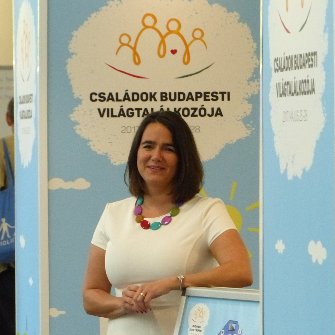 Katalin Novak standing in front of a booth. The booth is light blue and has small clouds on it. Just above her in the background the logo of the World Congress of Families in visible. Line drawing of two adults and two children, with the text "World Congress of Families in Budapest" under it. Novak is wearing a white T-shirt with a large necklace made out of large, colourful oval shaped beads.