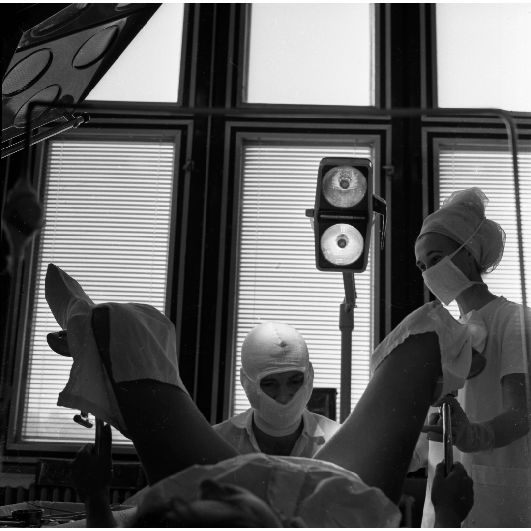 Black and white picture of a gynaecological examination. In the foreground a woman is lying down with her legs up on stilts. In the middle of the picture, a doctor is examining the patient. The doctor is wearing a white almost balaclava like headgear. A nurse stands to the right of the picture. In the background the windows provide some light alongside a standing light towards the centre of the picture, behind the doctor.