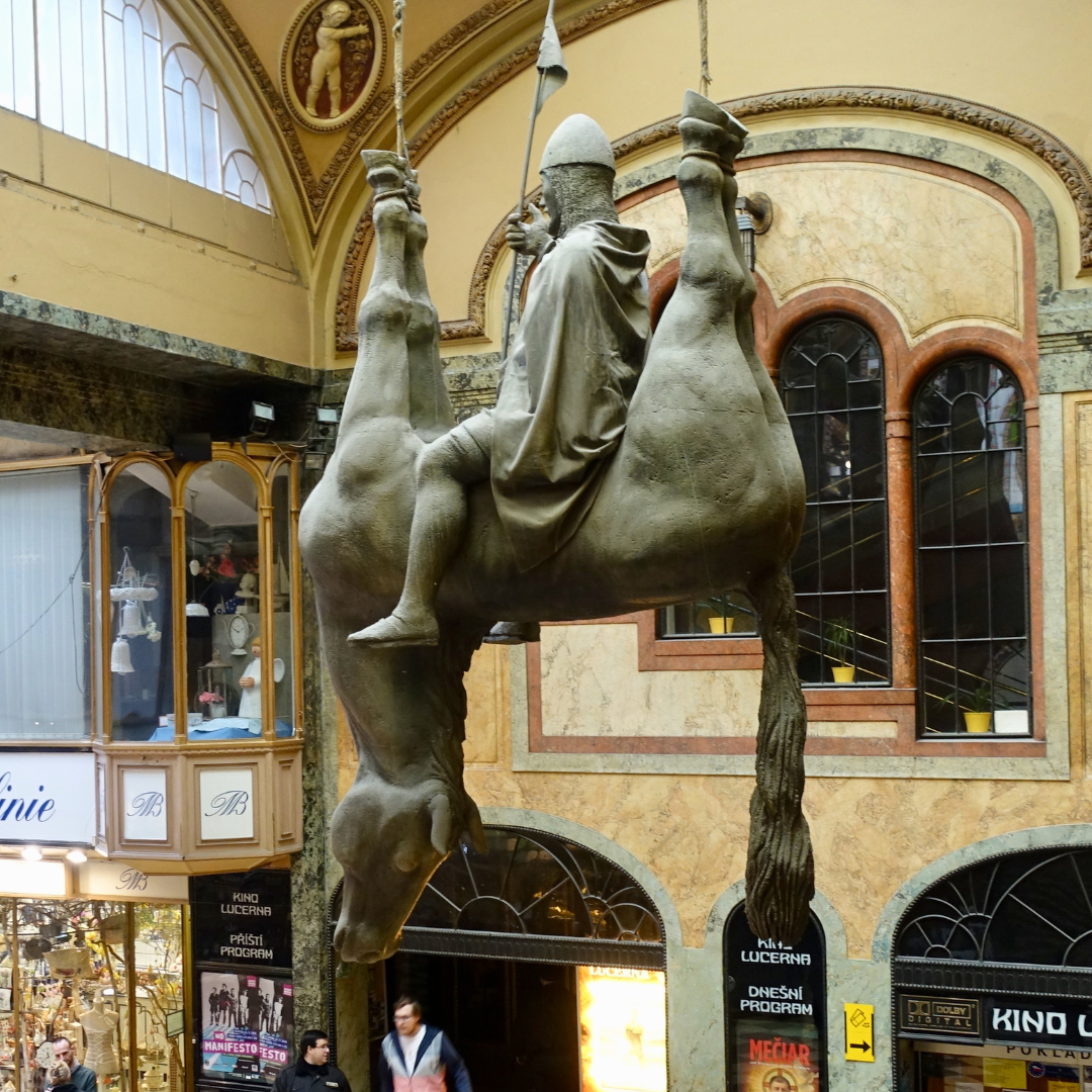 Statue of David Cerny in Prague. A statue of a horse is suspended from the ceiling by its legs, with a figure sitting on its belly. The figure is St Wenceslas. He is sitting straight on the hanging horse with a spear in his right hand. The statue is located inside a passage. The walls are painted yellow and they are adorned with various abstract motives. Below shopfronts are visible.