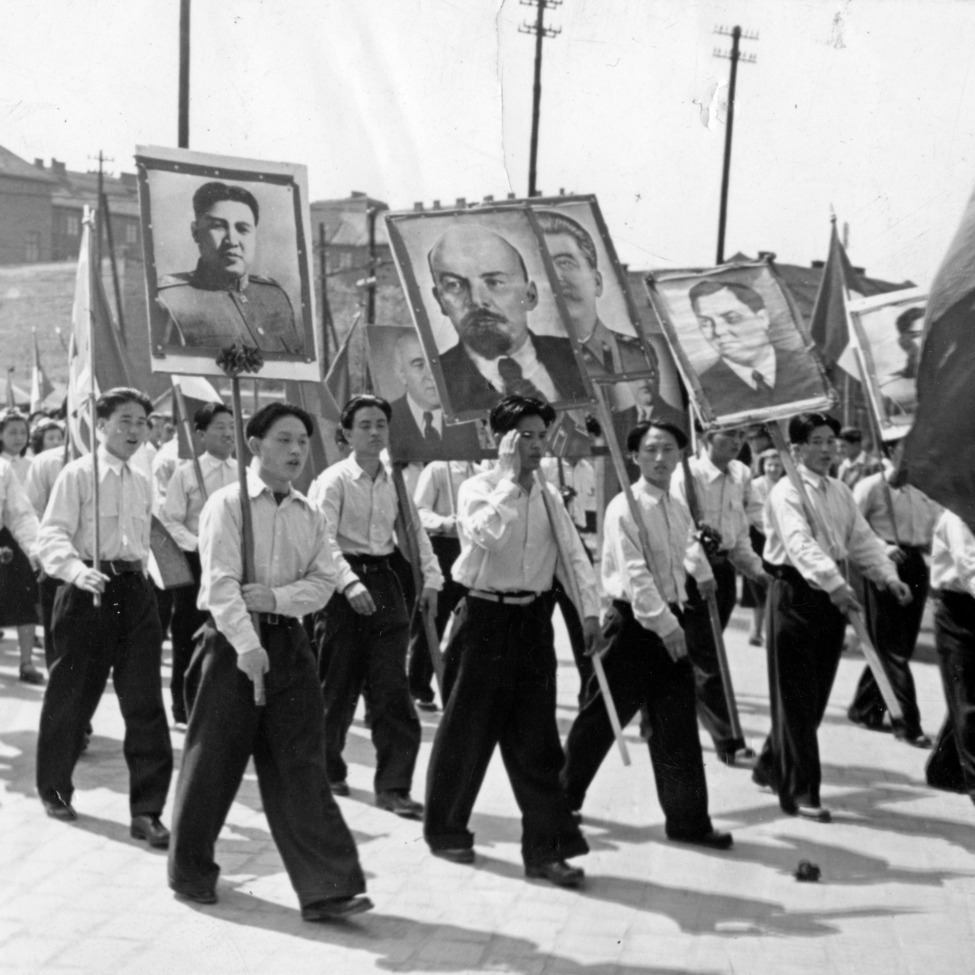 Black and white picture showing Korean students dressed in white shirts and black trousers during the 1 May celebrations. The front row is carrying placards of Lenin and other socialist leaders. The second row is carrying various flags. In the background you can see three lampposts and on the top left-hand corner some buildings of various heights.