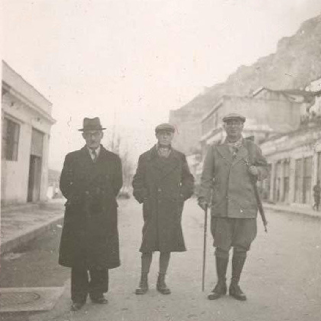 A somewhat faded sepia photograph with three man standing in the middle. The man on the left is wearing a long, stylish-looking black coat, a hat and black trousers. The man in the middle is wearing what looks like a long wool coat and a French cap. The man on the right is wearing a jacket, with trousers tucked into high boots. He has a riffle on his right side and he is holding a cane in his left hand. They are standing on a street with a big hill visible on the right hand side of the picture in the background. On their right and left, there are a row of low houses. Behind them on the street there are some trees in the distance.