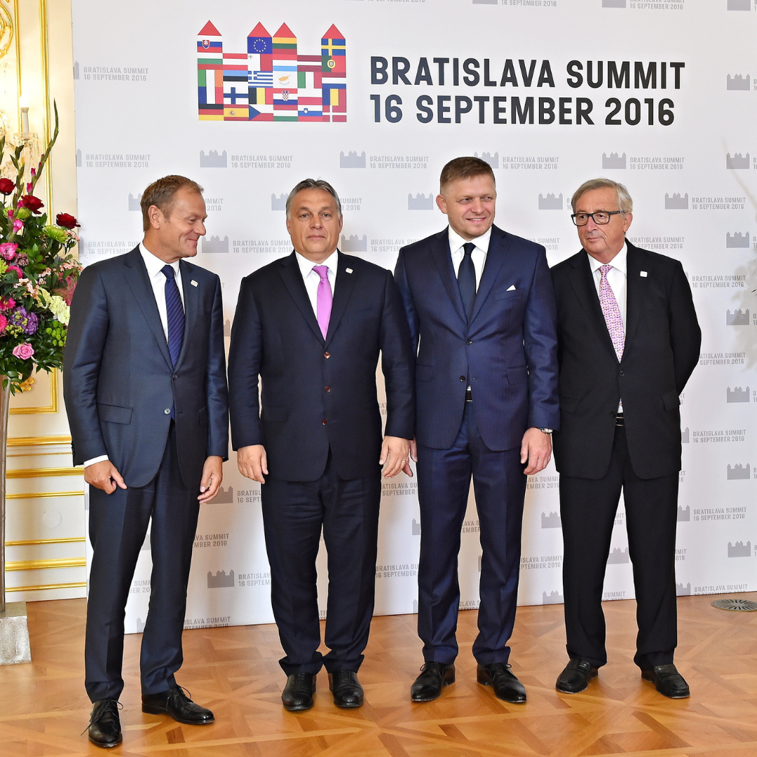 Four men, standing in front of a press wall. On the top of the wall on the left hand side there is a castle resembling Bratislava castle made up of EU flags. Next to the flag castle Bratislava Summit 16 September 2016 Is written. Donald Tusk on the left is standing next to Viktor Orban, Robert Fico and Jean-Claude Juncker.