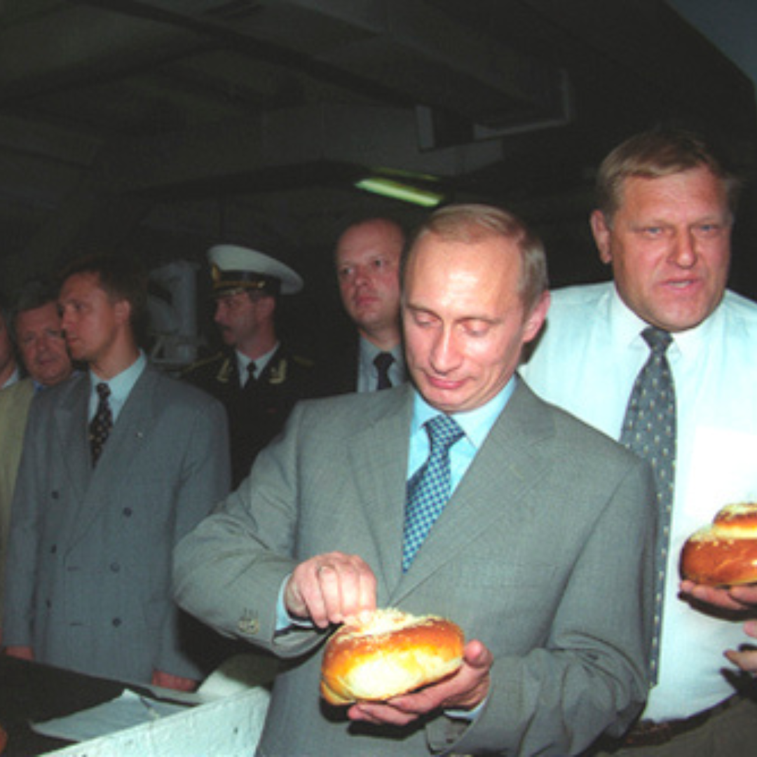 Putin standing with other people around him. He has a piece of pastry in his left hand and seems to be picking at it with his right hand. He is wearing. a grey suit with light blue shirt and a mid-blue tie.
