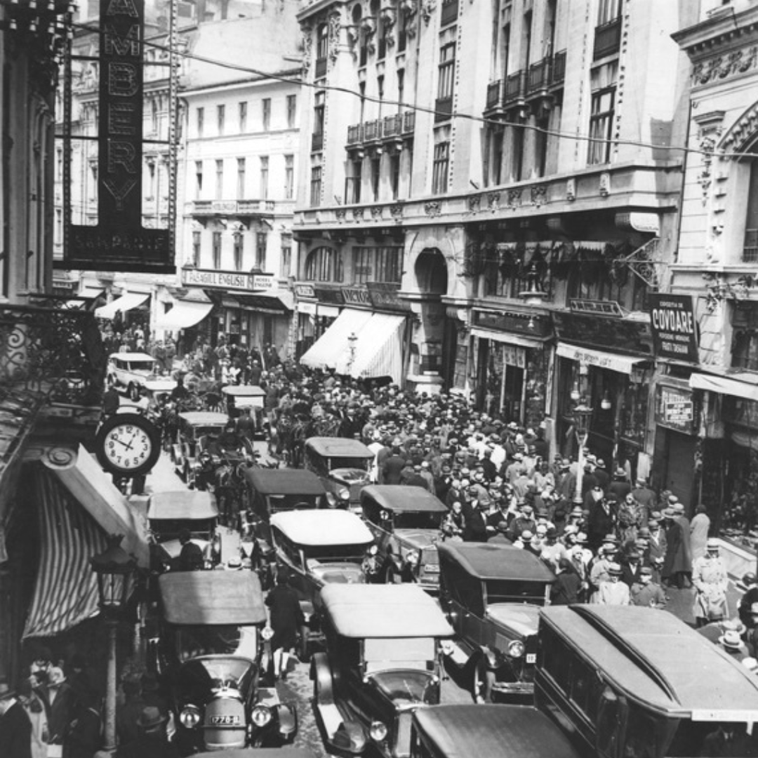Street scene. Cars are back to back on the road. The pavements are also full of people. The narrow street is surrendered by three and four storey buildings.