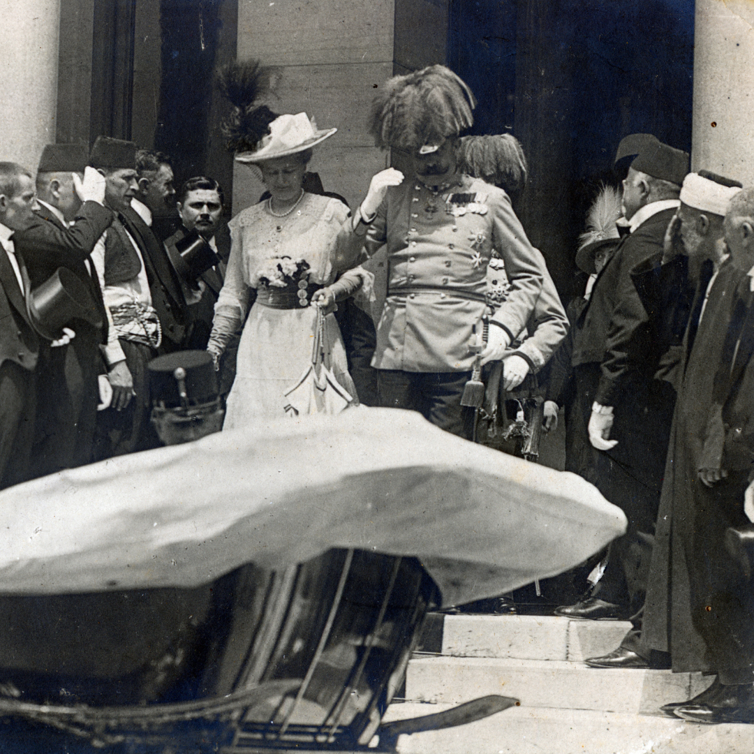Franz Ferdinand and his wife leaving the town hall just before his assassination. They are about to ascend a few steps to their carriage waiting for them below, part of the carriage is visible on the photo. They are surrounded by men, some of whom are giving them a military salute. Sophie is wearing a white dress and a white had with a feather. Franz Ferdinand is wearing a military uniform.