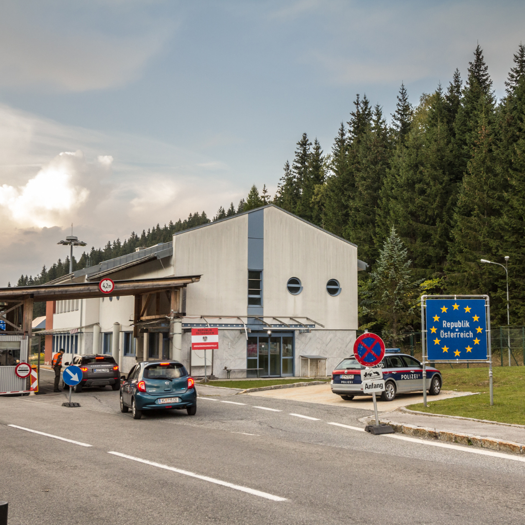 Cars pass through a border crossing station surrounded by forest, with signs indicating entry into the Republic of Austria and posted traffic regulations.