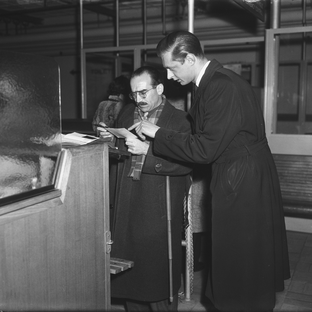 Two men standing at a service counter in an indoor public building; the man on the left, wearing glasses, a scarf, and an overcoat, reviews documents while the man on the right points to the papers.