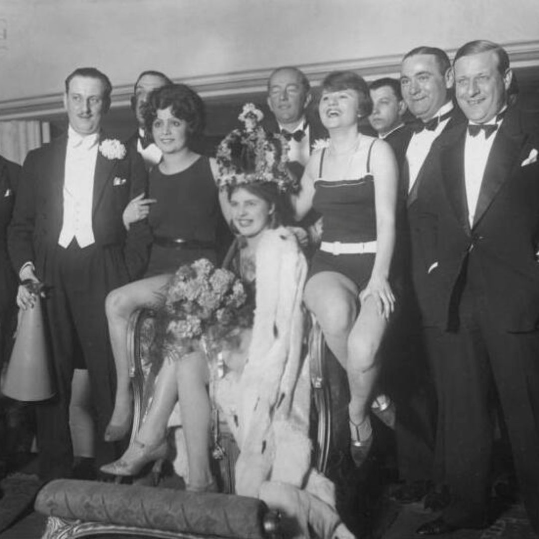 Black and white photograph from 1927 showing a group of elegantly dressed men and women at a beauty contest in Berlin. The women are wearing flapper-style dresses and accessories, while the men are in tuxedos, posing together and smiling for the camera.