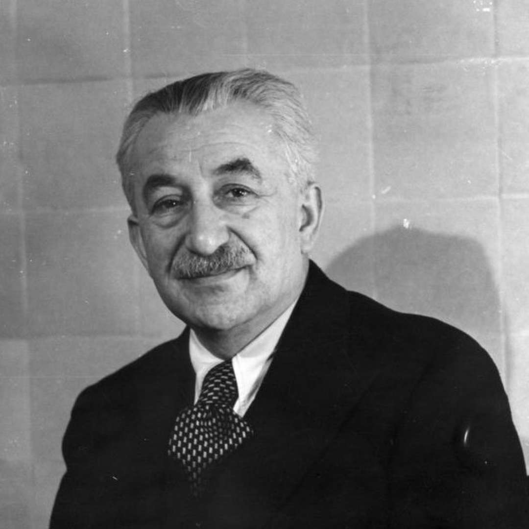 A black-and-white portrait of an older man with neatly combed gray hair and a mustache, wearing a dark suit, white shirt, and patterned tie. He is seated and smiling softly, looking slightly toward the camera. The background consists of plain, light-colored tiles.