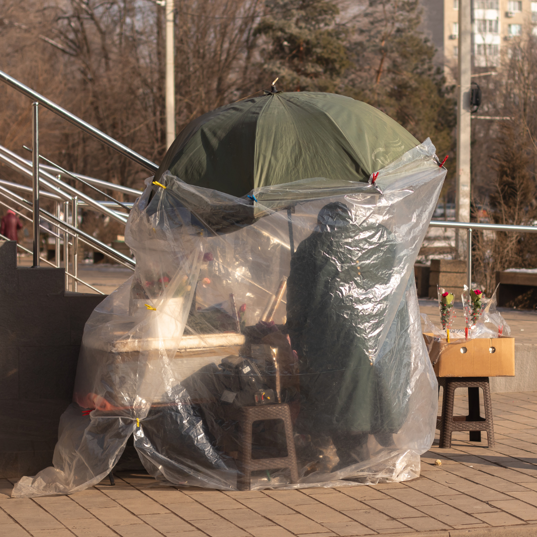 A street vendor during the Covid-19 pandemic sits under a makeshift shelter constructed from a large green umbrella and clear plastic sheeting, likely for protection. The vendor is surrounded by various items, possibly for sale, and is seated on a small stool. The scene is set outdoors in an urban area, with a staircase railing and trees visible in the background.