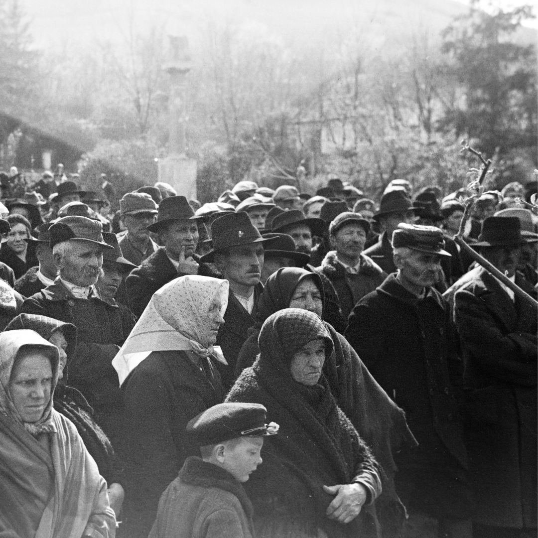 Black-and-white historical photograph of a large crowd gathered outdoors in a rural village setting. The crowd is made up mostly of peasants wearing heavy coats, hats, and headscarves, suggesting cold weather. Several women and elderly people stand in the foreground, along with a young boy in a cap. Faces appear serious and attentive, as if listening or waiting. Bare trees and simple buildings are visible in the background, reinforcing the rural, early-20th-century atmosphere.