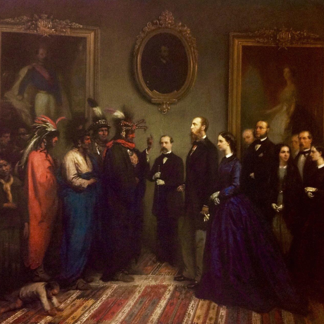 A 19th-century oil painting showing a formal meeting between two groups indoors. On the left, several Indigenous Kickapoo delegates stand together, wearing traditional clothing, feathered headdresses, and draped garments. On the right, European figures in dark formal attire—including a woman in a deep blue dress—stand facing them. The figures are arranged in a line as if greeting one another. Ornate framed portraits hang on the wall behind them, and a patterned rug covers the floor, emphasizing a ceremonial, diplomatic setting.