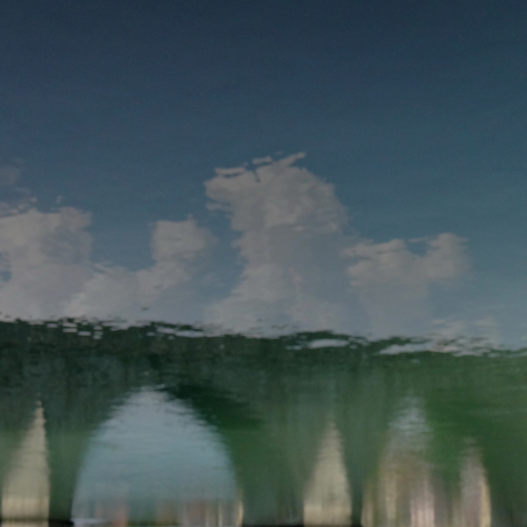 Reflection of the Mehmed Paša Sokolović Bridge shimmering in gently rippling water, its historic stone arches distorted into soft, wavy shapes beneath a blue sky with scattered clouds, with trees framing the scene.