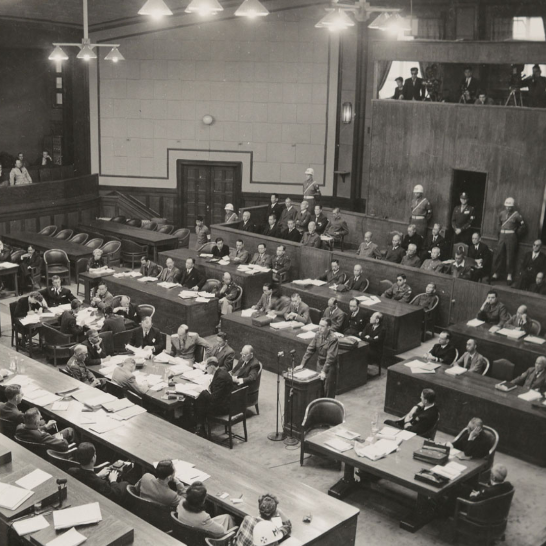 Black-and-white photograph of the courtroom of the International Military Tribunal for the Far East, with judges seated at raised benches, lawyers and officials at long tables, and a defendant speaking at a central podium while guards stand along the walls.