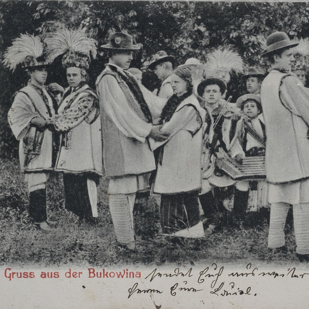 Black-and-white historical photograph of a group of men, women, and children standing outdoors, dressed in traditional folk clothing. Several individuals wear embroidered garments and tall feathered headdresses. In the center, a young couple faces each other holding hands, suggesting a dance or ceremonial moment. To the right, a small group of boys holds musical instruments, including what appears to be a violin and a cimbalom-like instrument. Trees form a leafy backdrop behind the group. At the bottom, printed in red, is the German phrase “Gruss aus der Bukowina” (“Greetings from Bukovina”), alongside a handwritten message in cursive.