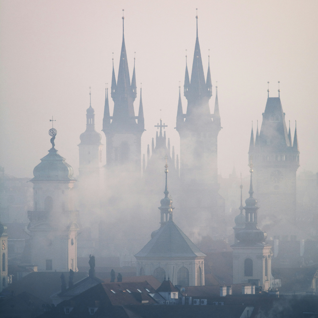 Misty cityscape of Prague with multiple historic church towers and spires rising above rooftops. The skyline is partially obscured by fog, creating a soft, atmospheric scene with pale light and muted tones. Distinctive Gothic towers and domes emerge through the haze, suggesting an early morning or winter setting.