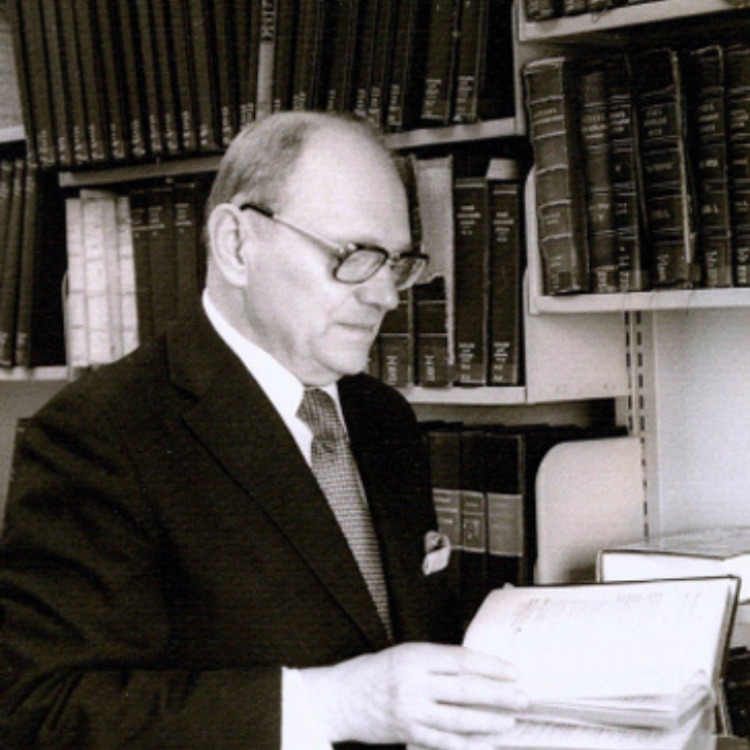 Black-and-white photograph of Omelian Pritsak standing in a library, wearing a suit and glasses, holding and examining a stack of papers. Behind him are shelves filled with neatly arranged books.