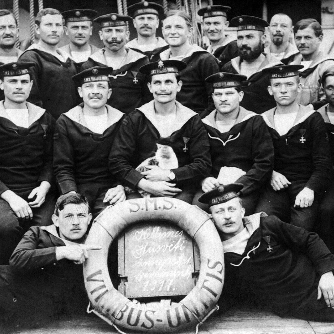 Black-and-white group portrait of sailors aboard the battleship SMS Viribus Unitis. Around fifteen crewmen in dark naval uniforms and caps pose closely together on deck, with ropes and ship rigging behind them. In the center, a seated sailor holds a small light-colored cat. A large life ring at the front is marked “S.M.S. VIRIBUS UNITIS,” with two sailors reclining on either side. The image has a formal yet relaxed feel, with several sailors looking directly at the camera.