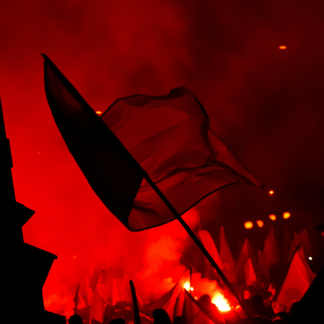 A nighttime demonstration bathed in intense red light from flares and smoke. A large flag on a pole waves prominently in the upper half of the frame, silhouetted against a glowing red haze. Below, a crowd of people is barely visible in silhouette, holding more flags and banners, with a bright burst of flame from a flare near the bottom center. The overall scene is dark and dramatic, dominated by deep reds and blacks.