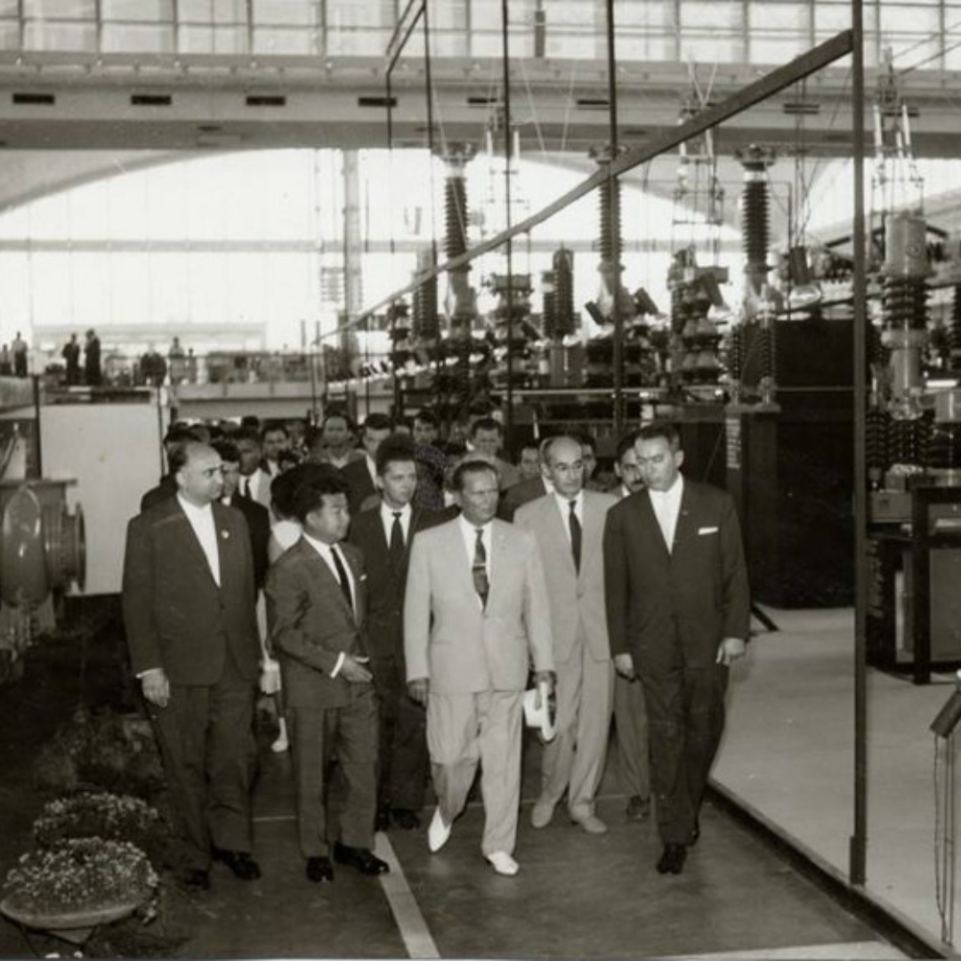 Black-and-white photograph of a group of officials walking through an Energoinvest industrial hall during a factory visit. At the centre, Emerik Blum and Yugoslav president Josip Broz Tito walk alongside members of a foreign delegation, all dressed in formal suits. They are touring a large electrical engineering facility filled with heavy industrial equipment, transformers, and high-voltage components.