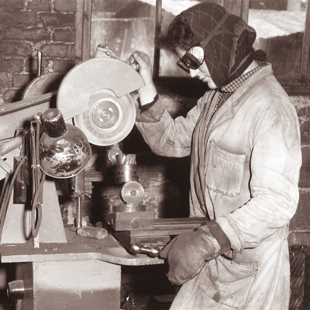Black-and-white archival photograph of a woman factory worker wearing a patterned headscarf, safety goggles pushed up on her forehead, and a heavy work coat with rolled sleeves. She stands at a metalworking machine (a bench grinder paired with a small lathe or precision vice setup), one gloved hand resting on the workpiece and the other raised near the grinding wheel. A gooseneck work lamp and hand tools are visible on the workbench in front of a brick wall.