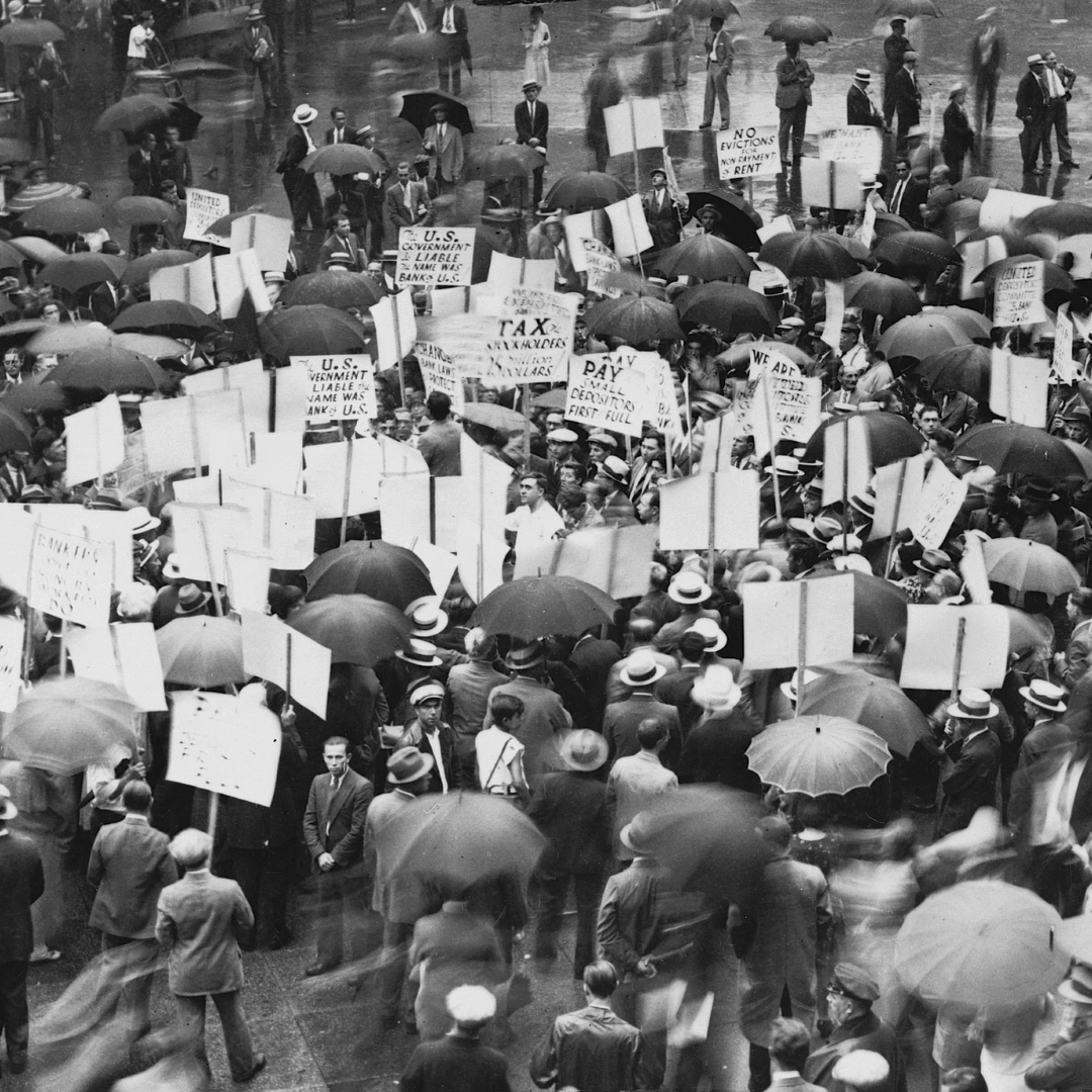Black-and-white photograph taken from above of a large crowd of depositors under umbrellas in the rain outside the failed Bank of United States in New York, around 1930–31. Many hold protest placards with slogans such as "Pay small depositors first in full," "Tax stockholders," "U.S. Government liable – name was bank's," and "No evictions for non-payment of rent."