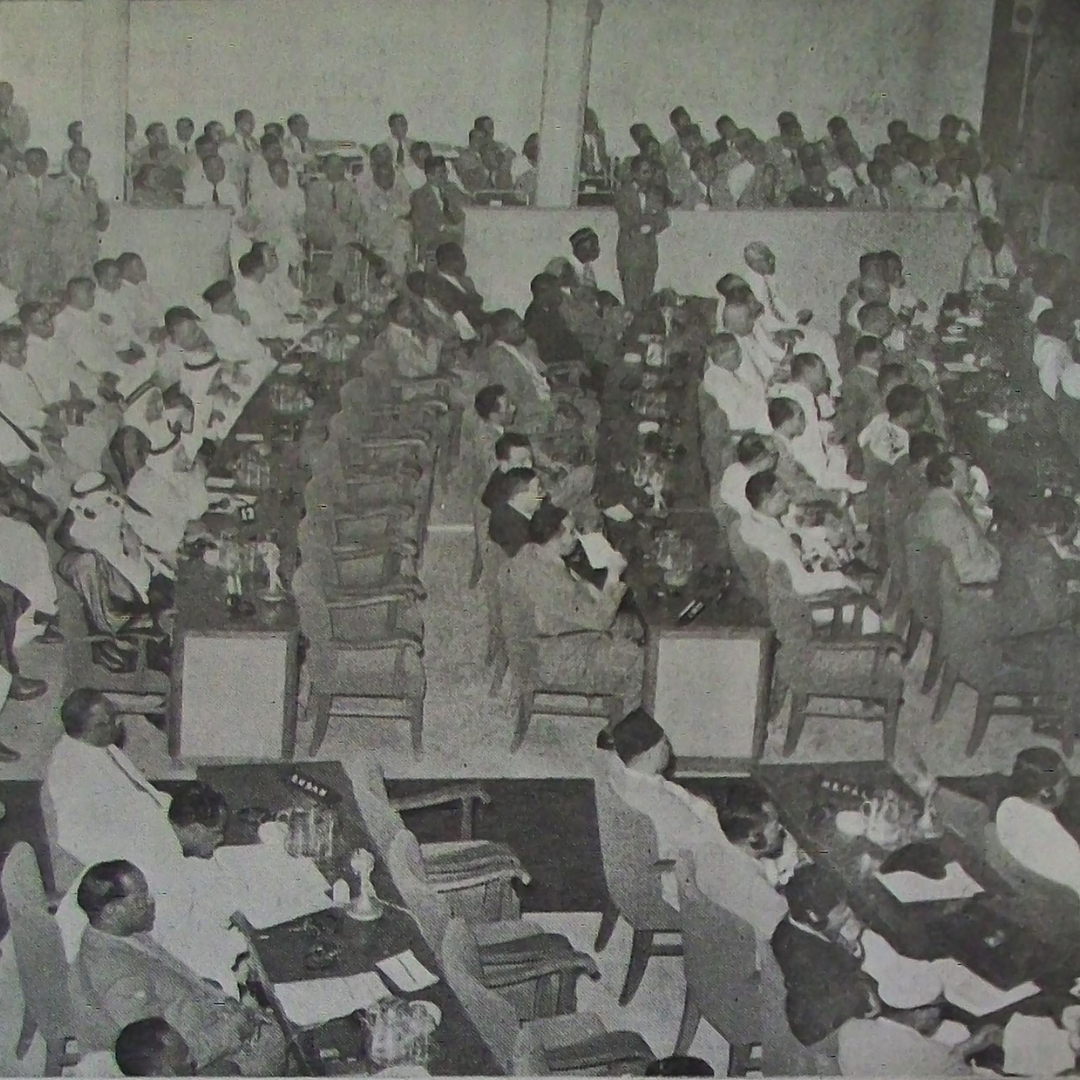 Black-and-white archival photograph of a plenary session at the Bandung Conference, showing delegates seated at long rows of tables on a conference floor, with an audience of observers seated in tiered galleries above.