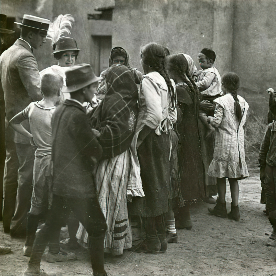 Black-and-white photo of a crowd of women and children in long skirts, headscarves, and shawls gathered on a dirt street beside a plain stucco wall, with a man in a straw boater and a woman in a feathered hat at the left.