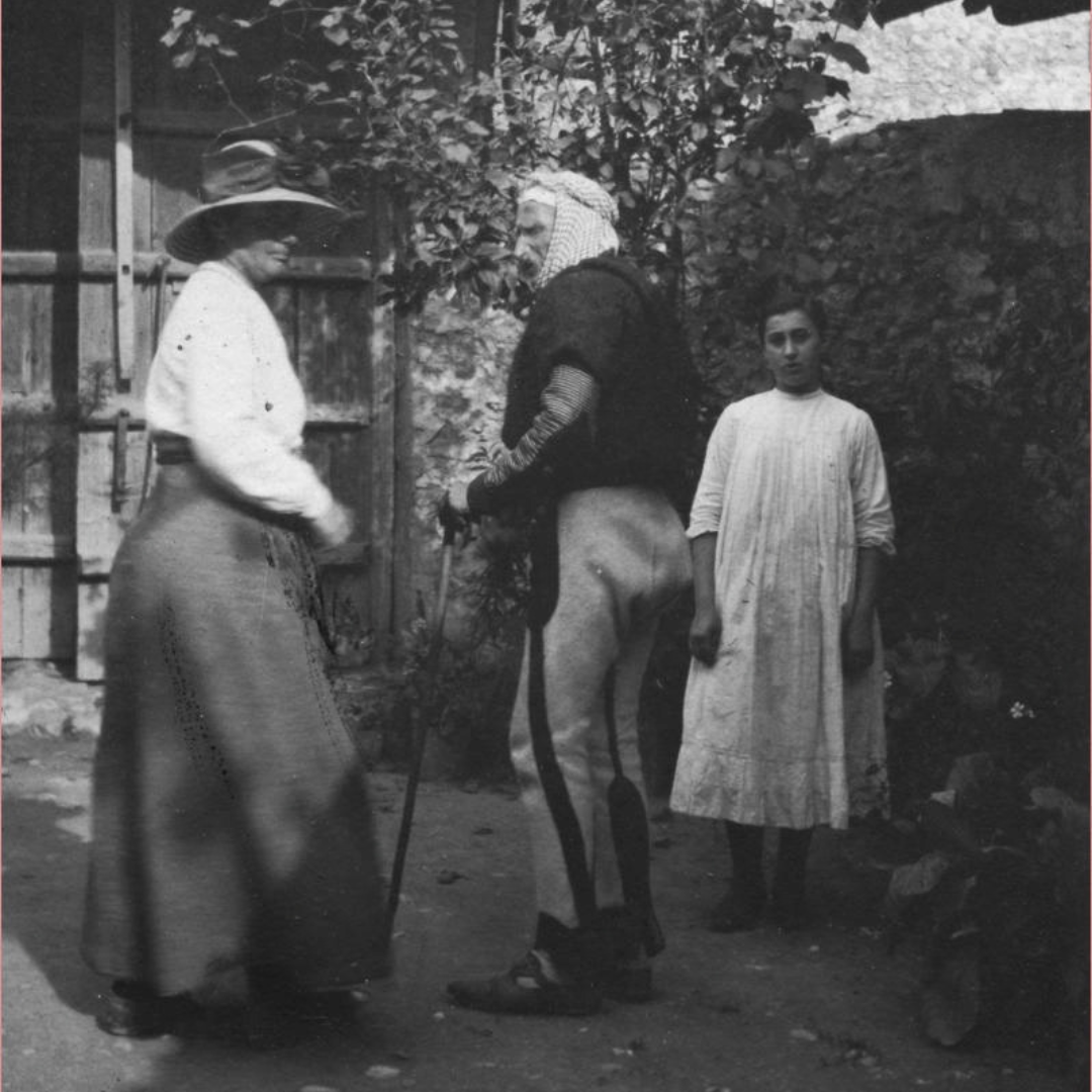 Edith Durham in a wide-brimmed hat and long skirt talking with the bearded bajraktar of Koplik, who leans on a stick, while a girl in a white dress watches, Shkodra, Albania, 1912.