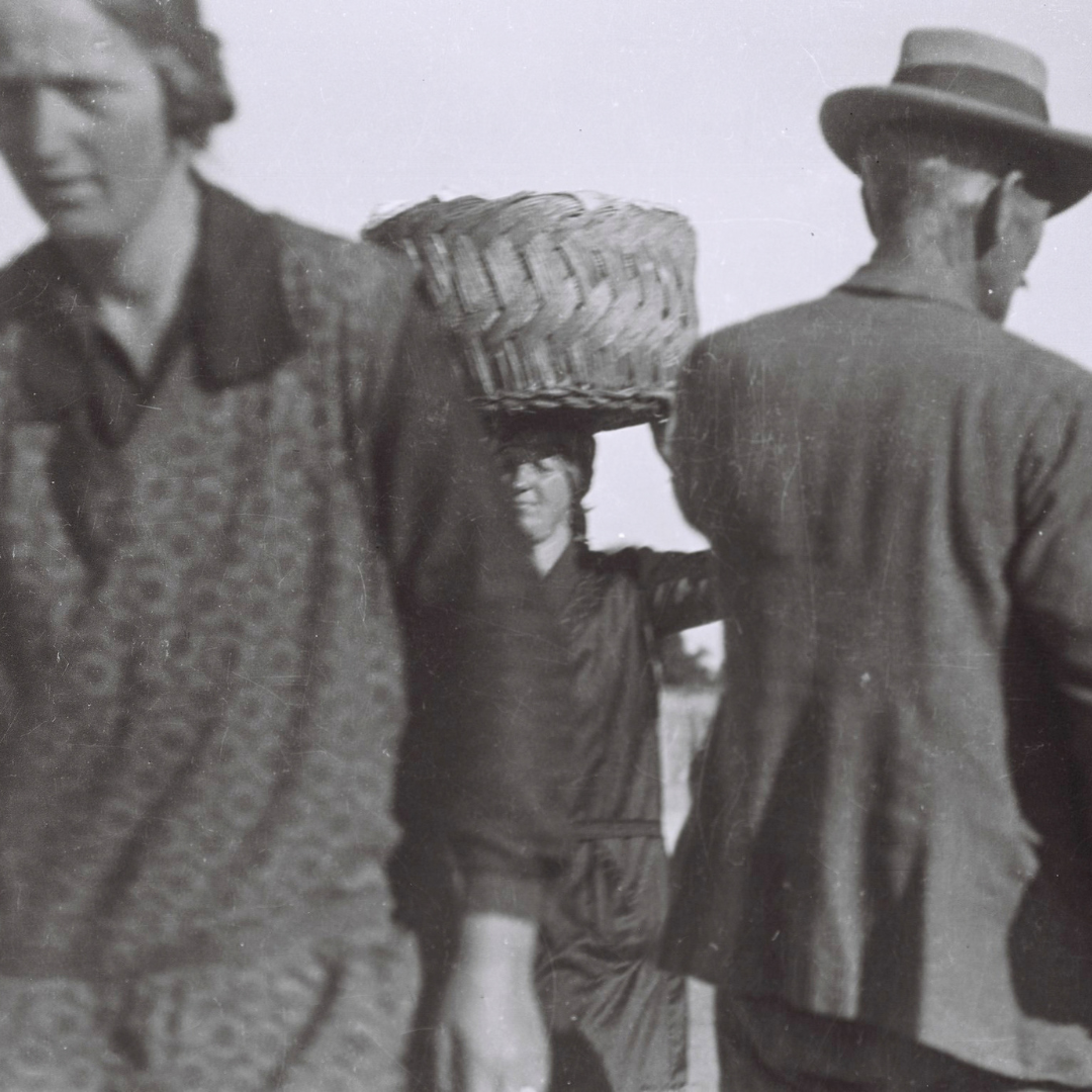 Black-and-white photograph of a woman carrying a large woven basket on her head, framed between a woman in the foreground and a man in a hat seen from behind.